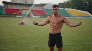 Vücut geliştiren biri futbol sahasında fıskiyenin su damlalarının altında durur ve kaslarını gösterir. Özgürlük kavramı. Yavaş çekim
