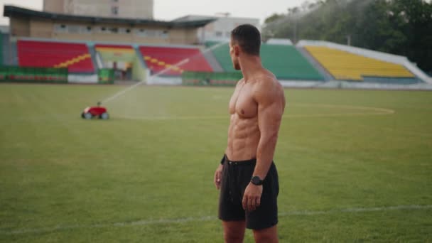En naken bål kroppsbyggare vilar efter träning på stadion och titta på sprinkler arbete på fotbollsplanen. Långsamma rörelser — Stockvideo