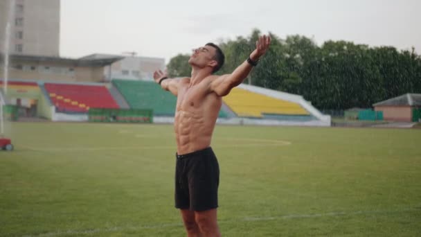 Muskularny mężczyzna stoi pod kroplami wody ze zraszacza na boisku piłkarskim z wyciągniętymi ramionami. Zwolnij trochę. Koncepcja wolności i sukcesu — Wideo stockowe