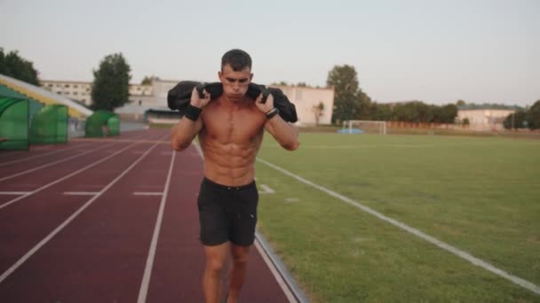 A naked torso bodybuilder runs along the treadmill of a city stadium holding a sandbag on his shoulders. Slow motion — Stock Video