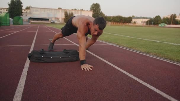 Bodybuilder training in het stadion en doet kern- en schouderkrachtoefeningen met een zandzak terwijl u op een plank staat — Stockvideo