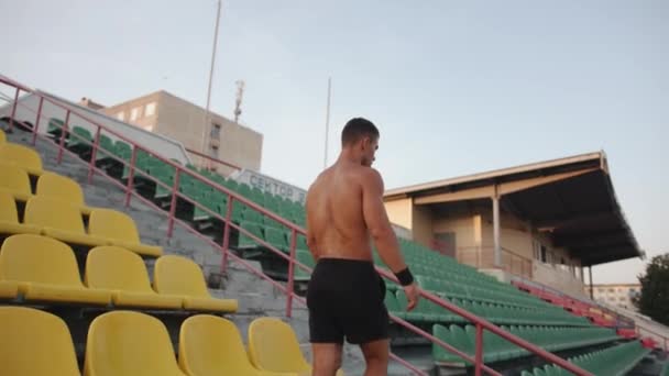 Навчання м'язистого чоловіка на стадіоні і під час його відпочинку стоїть на тлі пластикових сидінь трибуни. Повільний рух — стокове відео