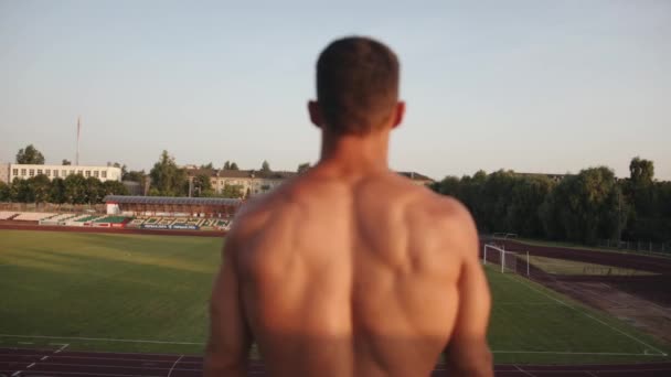 Un giovane uomo muscoloso salta sul posto sullo stadio e flette i muscoli della schiena. Vista posteriore. Rallentatore — Video Stock