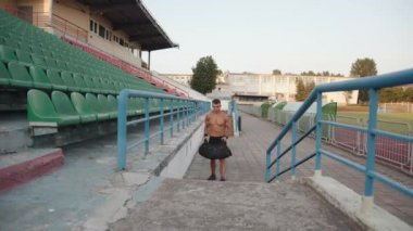 Bir vücut geliştirme ustası stadyumdaki tribünün merdivenlerine tırmanıyor ve antrenman için omuzlarına kum torbası takıyor.