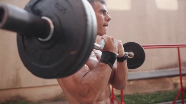 Hombre musculoso joven entrenando en un campo de deportes abierto y bombea sus bíceps y tríceps con una barra en forma de w. Movimiento lento — Vídeo de stock