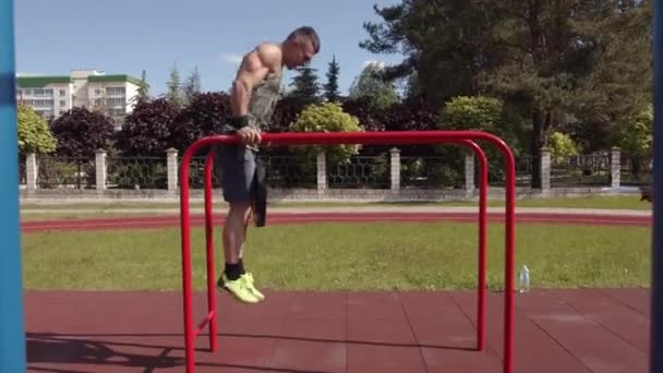 Homem musculoso treinando no campo esportivo e fazendo flexões nas barras irregulares com um disco da barra no cinto — Vídeo de Stock