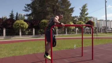 Spor sahasında kaslı bir adam antrenman yapıyor ve dengesiz barlarda halterden kopmuş bir diskle şınav çekiyor.