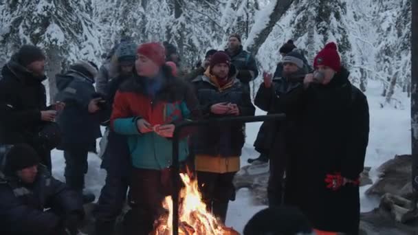 Regio Moermansk, Rusland - 10 januari 2021: Een groep toeristen tijdens een winterwandeling zit rond een kampvuur thee te drinken en sandwiches te eten — Stockvideo