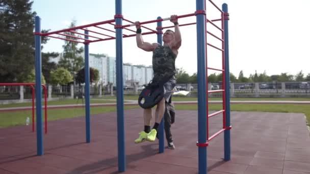 Minsk, Belarus - July 10, 2021: A bodybuilder pulls himself up on a horizontal bar with a disc from a barbell on his belt and another man records the result in a notebook — Stock Video