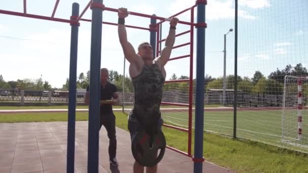 Minsk, Belarus - July 10, 2021: A muscular man pulls up on the horizontal bar with a disc from a barbell on his belt and another bodybuilder watches the exercise — ストック動画