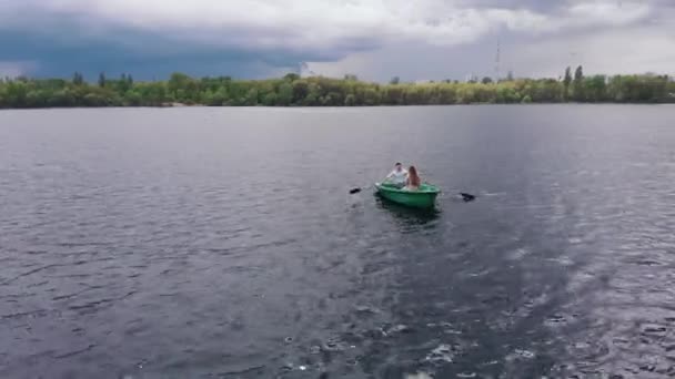 Légi kilátás egy fiatal lány és egy férfi egy evezős hajón a folyón, közel a mólóhoz és a kilátás a városra a távolban — Stock videók