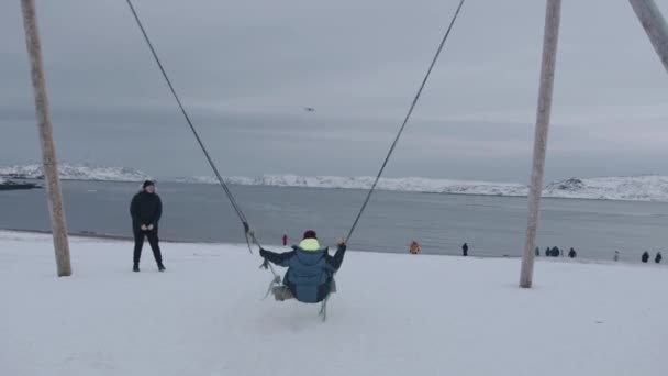 Região de Murmansk, Rússia - 10 de janeiro de 2021: Uma jovem cavalga em uma corda balançando em uma colina nevada junto ao mar com turistas ambulantes. Movimento lento — Vídeo de Stock