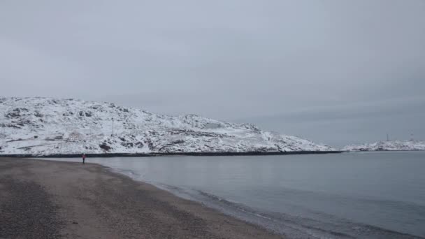 Rustige winterzee met golven op het zand en een silhouet van een vrouw tegen de achtergrond van besneeuwde bergen. Eenzaamheid en reflectie — Stockvideo