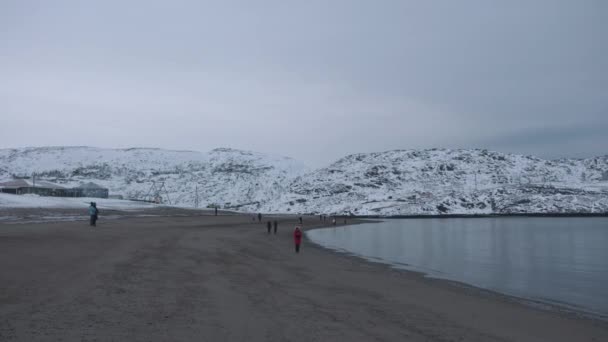 Toeristen wandelen langs de zeekust van de baai omgeven door sneeuwruggen en nederzetting aan de kust — Stockvideo