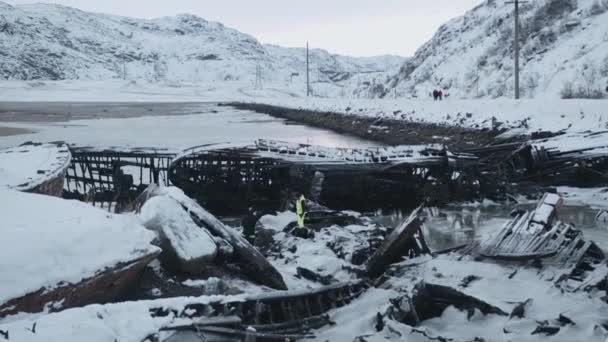 Teriberka köyü yakınlarındaki kış denizi kıyısında terk edilmiş gemilerin iskeletleri ve kıyı boyunca yürüyen bir grup insanın görüntüsü. — Stok video