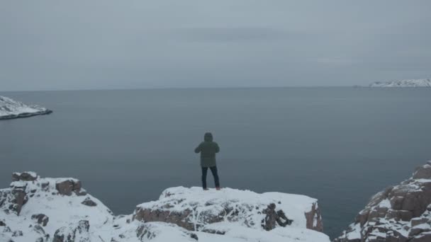 Un hombre se para en una roca y mira a la bahía de mar rodeada de crestas cubiertas de nieve. Concepto de soledad y reflexión. Vista trasera — Vídeos de Stock