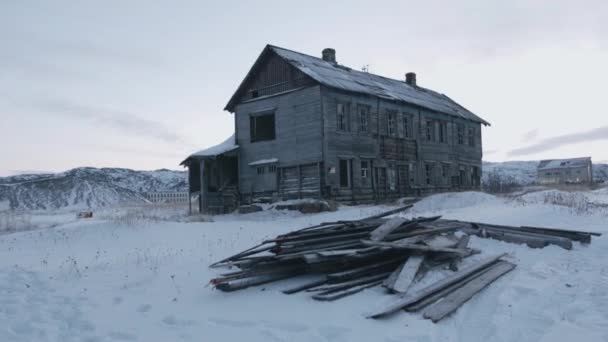 Old dilapidated wooden house with broken windows on a background of snowy rocks and a dump of boards in a vacant lot — Stock Video