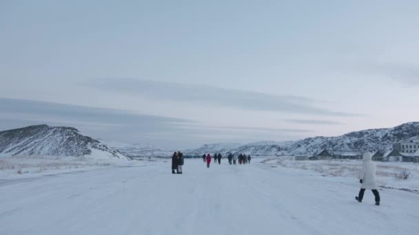 Een groep toeristen loopt langs een brede besneeuwde weg langs de straat van het dorp Teriberka. Achteraanzicht — Stockvideo