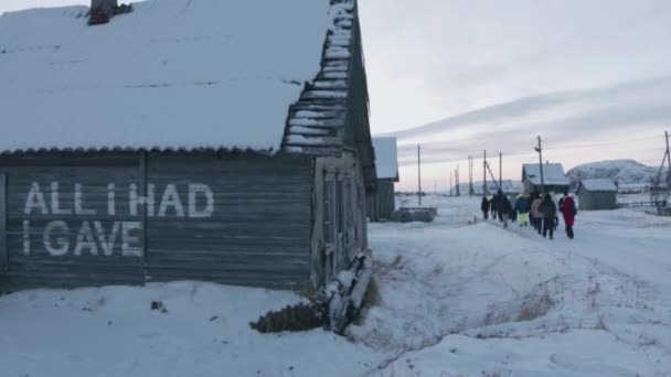 La gente in escursioni va lungo la strada del villaggio di Teriberka passato vecchie e distrutte case di legno in una giornata d'inverno. Vista posteriore — Video Stock