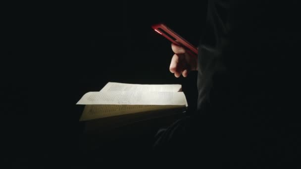 एक माणूस अंधारात पुस्तक वाचतो आणि पृष्ठांवर मोबाइल फोन फ्लॅशलाइट चमकतो. बंद करा — स्टॉक व्हिडिओ
