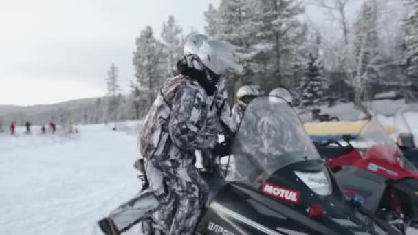 Regio Moermansk, Rusland - 10 januari 2021: Een man stapt met een vrouw-passagier achter het stuur van een sneeuwscooter en bereidt zich voor op een ritje samen met andere — Stockvideo