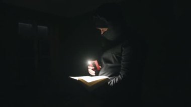 Kapüşonlu genç bir adam karanlık bir odada duruyor ve telefondan el feneri tutarak kitap okuyor.