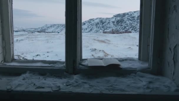 Kniha leží na starém okně bez skla opuštěného domu s výhledem na zasněžené skály a vítr otáčí stránky — Stock video