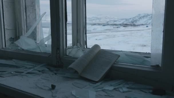 Primer plano de un libro sobre una ventana con fragmentos de vidrio en una antigua casa abandonada con vistas a rocas nevadas — Vídeos de Stock