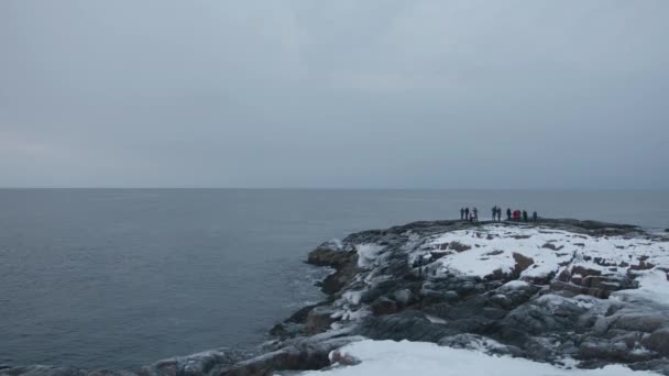 観光客のグループは雪の丘に沿って歩き、海の波が石に対してどのように破るかを賞賛します。 — ストック動画