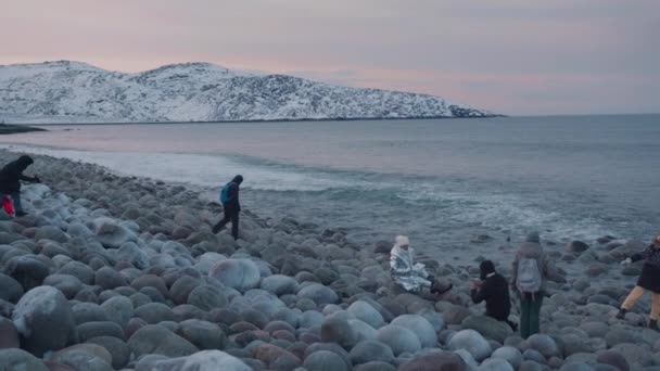 Region Murmansk, Russland - 10. Januar 2021: Touristen gehen und sitzen auf runden Felsbrocken am Strand von Dracheneiern in der Barentssee bei einem Winterausflug — Stockvideo