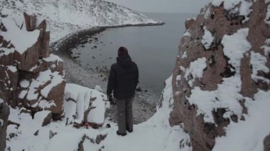 Genç bir adam uçurumun kenarında duruyor ve Barents Denizi kıyısındaki ejderha yumurtaları plajının manzarasına hayran. Arka plan