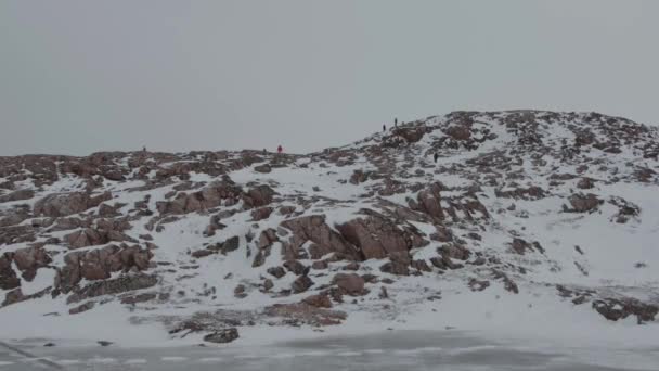 Un grupo de turistas trepan y caminan por la cima de rocas cubiertas de nieve con un reservorio congelado a los pies — Vídeo de stock