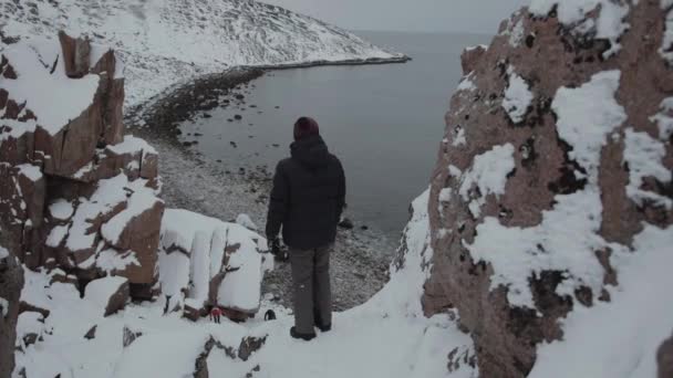 Een jonge man staat in een klif kloof en bewondert het uitzicht op het drakeneieren strand aan de kust van de Barentszzee. Achteraanzicht — Stockvideo