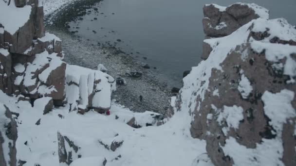 Vista superior de la costa del mar rodeado de rocas nevadas y un hombre a los pies hace su camino a través de rocas redondas — Vídeo de stock
