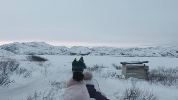 सूर्यास्त पर बर्फीली चट्टानों की पृष्ठभूमि पर पर्यटकों के साथ sleighs के साथ स्नोमोबाइल का स्तम्भ। धीमी गति। हाथ में पीओवी शॉट — स्टॉक वीडियो