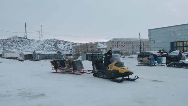 Murmansk περιοχή, Ρωσία - 10 Ιανουαρίου 2021: Ένας άνθρωπος πίσω από το τιμόνι ενός snowmobile με ένα έλκηθρο συνδέεται βόλτες έξω από το πάρκινγκ — Αρχείο Βίντεο