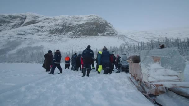 Region Murmansk, Russland - 10. Januar 2021: Vor der Kulisse eines verschneiten Waldes am Fuße des Berges spaziert eine Gruppe Touristen in der Nähe von Motorschlitten mit Schlitten. Zeitlupe — Stockvideo