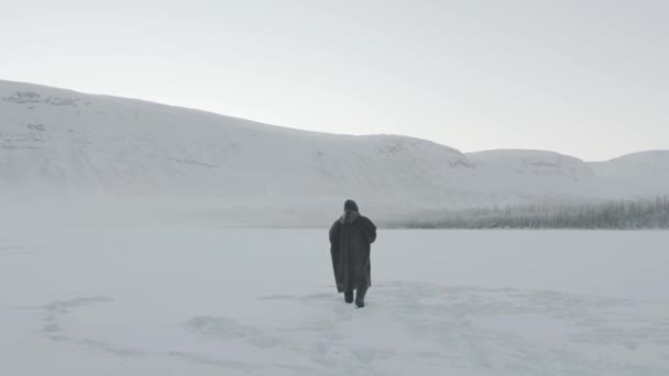 Een man in een brede jas met capuchon gaat langs een besneeuwd veld tegen de achtergrond van een berg. Achteraanzicht. Langzame beweging. Eenzaamheid concept. Privacy. Verlaten. Depressie — Stockvideo