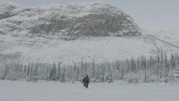 Een man in een met sneeuw bedekte jas en een beschermend masker gaat door de sneeuw tegen de achtergrond van een bos en een berg. Eenzaamheid concept. Privacy. Verlaten. Depressie. Spiritueel — Stockvideo