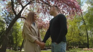 Güzel bir genç kız parktaki çiçek açan bir ağacın altında dururken erkeğiyle konuşuyor ve elini tutuyor. Yavaş çekim