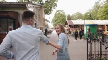 Genç bir kız erkeğin elini tutuyor ve onu parktaki alışveriş galerisinde yürümeye davet ediyor. Arkadan bak. Yavaş çekim