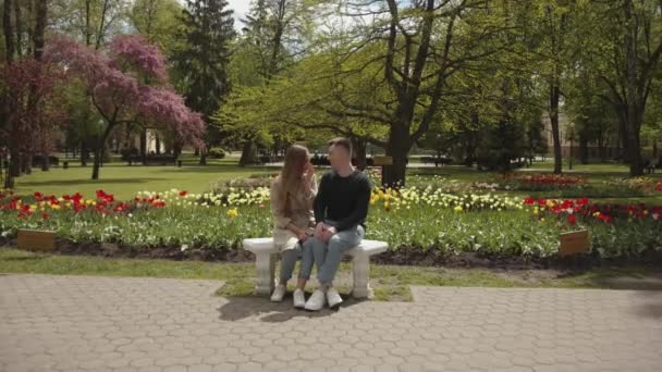 Pasangan kekasih yang menawan duduk di bangku dan berpegangan tangan melawan latar belakang tempat tidur bunga di taman. Gerakan lambat — Stok Video