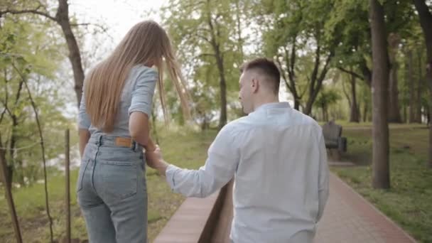 Een jong meisje gaat langs de stoeprand van het pad in het park en houdt zich vast aan de arm van haar geliefde man. Achteraanzicht. Langzame beweging — Stockvideo