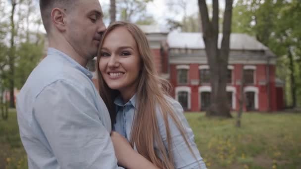 Krásný mladý pár stojí v parku na pozadí červené budovy a objímají se do kamery. Zpomalený pohyb — Stock video
