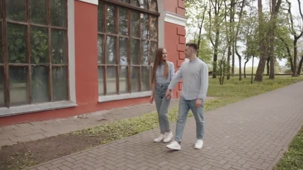 Et ungt par i kærlighed holder hinanden i hånden og går langs stien i parken forbi bygningen med store vinduer. Langsom bevægelse – Stock-video