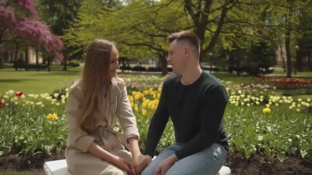 Um belo casal jovem está falando e rindo enquanto se senta em um banco contra o pano de fundo de um parque florescente. Movimento lento — Vídeo de Stock