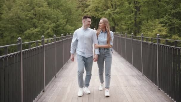 Una joven pareja enamorada va a lo largo del puente sobre el río en el parque en un día de verano y sonriendo mirándose. Movimiento lento — Vídeo de stock