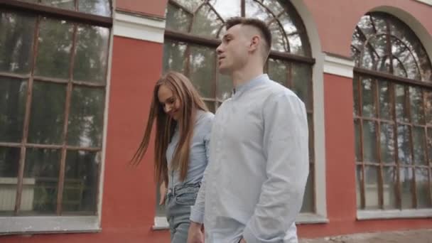 Um jovem casal apaixonado passa por um prédio com grandes janelas arqueadas e fala olhando um para o outro. Movimento lento — Vídeo de Stock