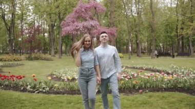 Genç romantik çift el ele tutuşup parktaki çiçeklerin arka planına ters düşüyor. Aile ve aşk kavramı. Yavaş çekim