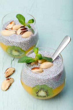 Sağlıklı kahvaltı. Cam bardakta mangolu Chia pudingi, mango püresi, kivi, Brezilya fındığı ve mavi ahşap arka planda nane şekeri. Seçici odak